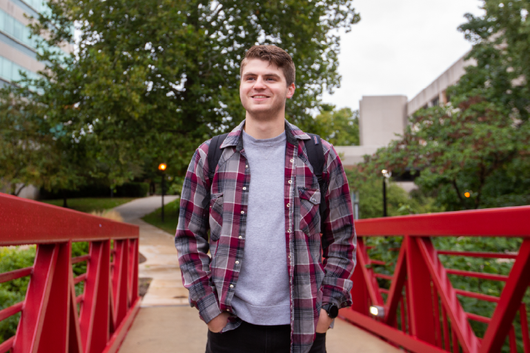 A male student wearing a backpack walks across a red metal bridge on the IU South Bend campus.